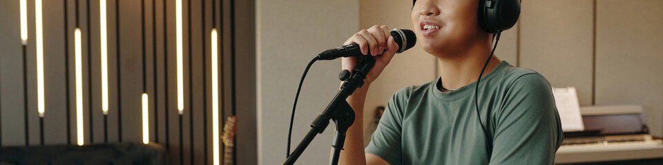 Young adult wearing headphones and singing into a microphone in soundproof recording studio with modern design and piano visible in background