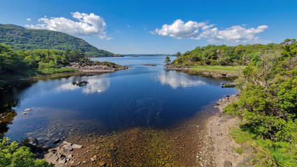 Obraz premium A breathtaking view of Muckross Lake under the clear blue sky, where the shimmering waters mirror the lush greenery and surrounding mountains, inviting all to embrace the beauty of nature.