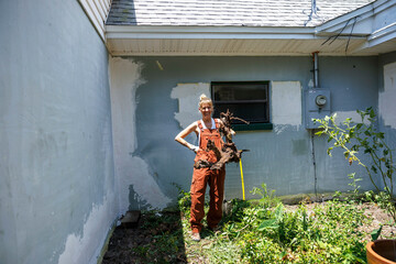 A smiling woman stands proudly next to a freshly dug spot in a garden, holding a shovel and a root clump, wearing orange overalls, Florida, USA
