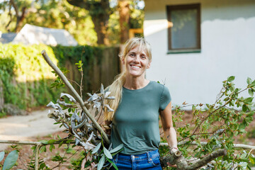 Smiling woman with blond hair holding cut branches while doing yard work outside a house, Florida, USA