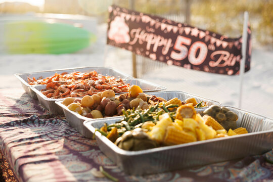 Outdoor picnic table decorated for a birthday celebration with a 'Happy 50th Birthday' banner and trays of delicious food in the warm sunlight, Florida, USA