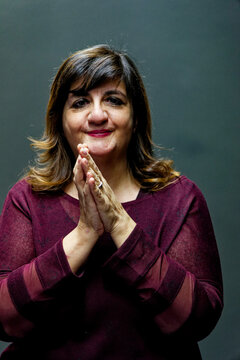 Smiling woman with hands clasped in a gesture of gratitude or request wearing a maroon sweater against a dark backdrop.