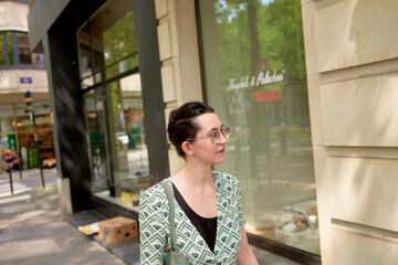 A woman in a patterned green cardigan walks by a city storefront with trees and reflections in the window.
