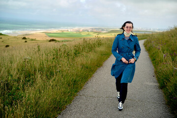 Woman in blue coat smiling while walking on a countryside path with a scenic coastline in the background