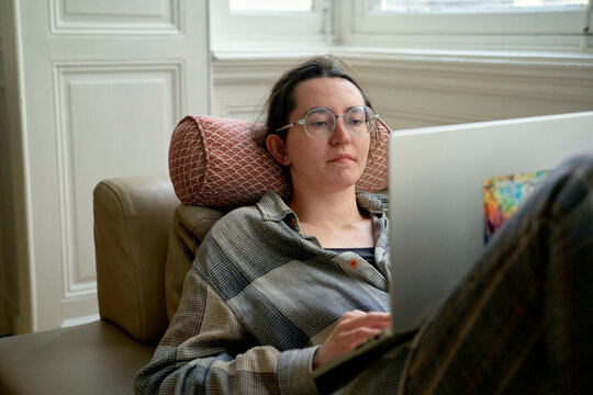 A focused adult wearing glasses and a checkered shirt is working on a laptop while reclining comfortably on a beige armchair.