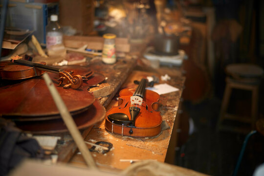 A violin and bow resting on a cluttered workbench in a violin maker's workshop surrounded by tools and wood shavings.