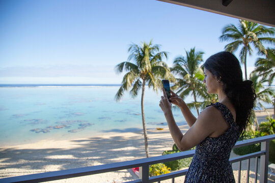 Woman takes a photo of a tropical beach view from a balcony with her smartphone, Titikaveka, Rarotonga, Cook Islands
