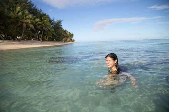 Woman smiling while wading in clear tropical waters with palm trees lining the beach in the background, Titikaveka, Rarotonga, Cook Islands