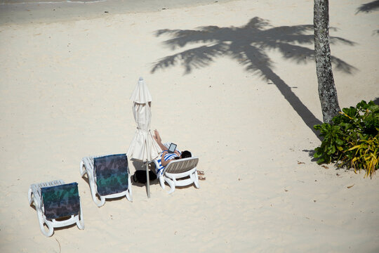 Sunbather relaxing  with an e-book under palm tree shadows on a sandy beach with an open white umbrella and lounge chairs, Titikaveka, Rarotonga, Cook Islands
