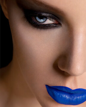 Close-up portrait of a woman with striking blue lipstick and dramatic eye makeup against a black background.