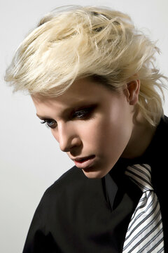 Side profile of a blond person with dramatic makeup wearing a black blazer and striped tie, looking downwards against a light background.