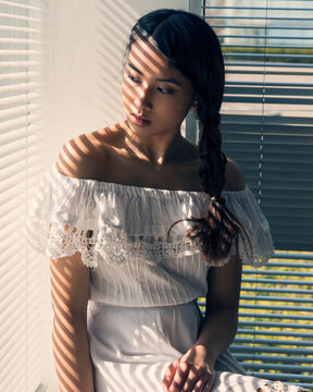 Woman in white off-shoulder dress standing by a window with sunlight casting striped shadows on her face and body.