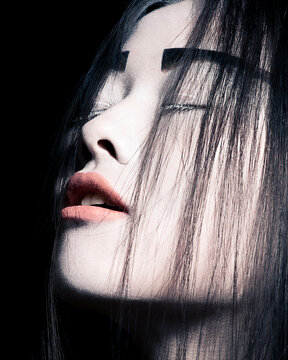 Close-up of a woman with dramatic makeup and striking features, her eyes closed and hair partially covering her face.