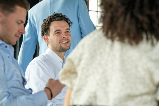 Smiling man in a blue shirt engaged in a conversation at a group meeting with partial views of other attendees.