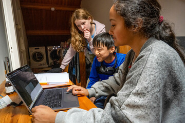 Three children are gathered around a laptop with one of them typing, giving the impression of a collaborative homework session in a home environment.