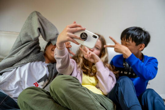 Three kids take a selfie together, one covering their face with a hoodie, another making a peace sign, and the third smiling at the camera.