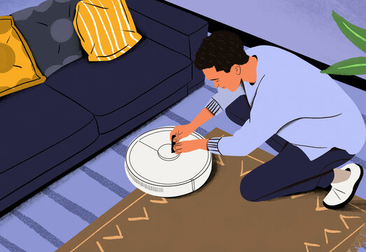 Man kneeling on a carpet to repair or set up a robotic vacuum cleaner next to a couch with a striped pillow.