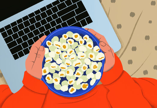 Overhead view of a person in an orange sweatshirt using a laptop and holding a blue plate of popcorn.
