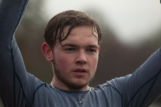 Young male athlete resting with hands on head after a strenuous outdoor workout, perspiration visible on his face, looking slightly away from the camera.