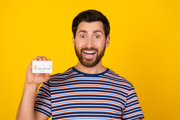 Portrait of impressed guy with bristle wear striped t-shirt presenting debit card astonished staring isolated on yellow color background