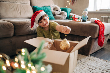 Kid boy decorating Christmas tree put off balls from the box authentic real celebration image...