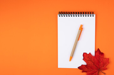 Flatlay composition with blank notepad, pen and autumn leaf on orange background. Top view. Copy...