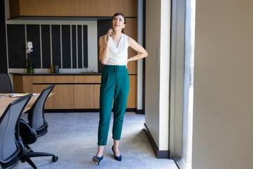 Talking on smartphone, businesswoman standing in modern office near window