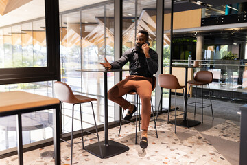 Fototapeta premium Talking on smartphone, African american businessman sitting at high table in modern office space