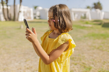 Girl 7 years old in profile in yellow dress takes photos on phone on lawn with palm trees in summer day