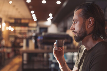 Pensive bearded hipster guy enjoying morning coffee for breakfast looking away in cafeteria, thoughtful caucasian male in casual outfit drinking beverage during break in cafe interior