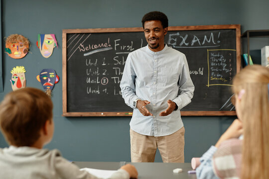 Medium full shot of smiling African American male teacher standing in front of young students in classroom wishing children good luck in exam, copy space