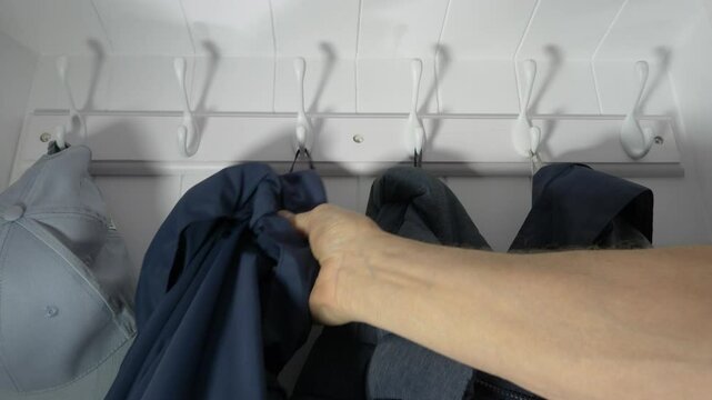 Closeup of a hand hanging a blue jacket on a white hook in a white cupboard, next to other clothing.