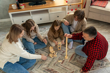 Happy family are sitting on floor, playing board game with wooden tower together at home while spending leisure time at cozy home during Christmas holidays. Indoor activities.