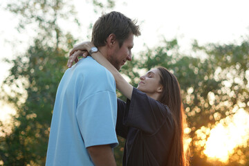 woman in a black shirt embraces a man in a blue shirt as they stand in a forest. The sun shines behind them, creating a warm, golden glow