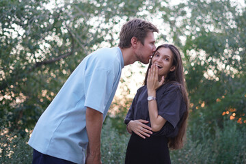 Fototapeta premium couple shares secret to each other's ear as they stand in a park, surrounded by greenery