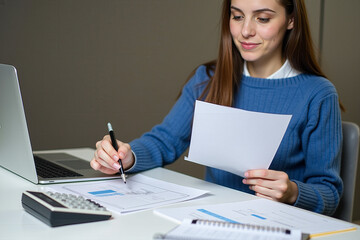 A young professional reviewing documents while working on a laptop, with a calculator and pencil on the desk. The setting suggests a modern office or home workspace, focusing on concentration and prod