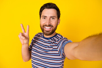 Portrait of optimistic guy with bristle wear striped t-shirt making selfie showing v-sign symbol isolated on yellow color background