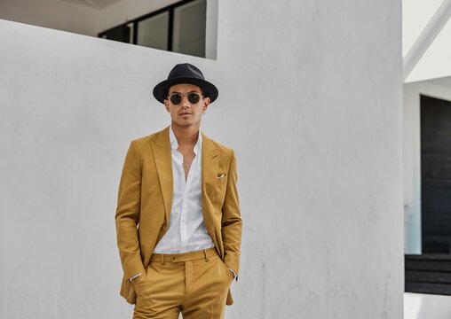 Urban Dapper: A stylish Asian man in a yellow suit, fedora, and sunglasses exudes confidence against a modern architectural backdrop. 