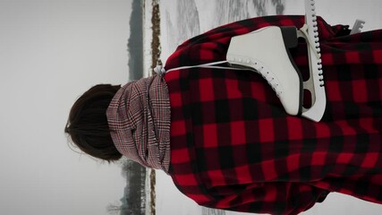 Back shot of woman with skates on shoulder walking along ice lake.Vertical video - Powered by Adobe