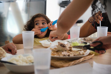 Mother is serving her daughter lunch at their kitchen table, while the rest of the family eats
