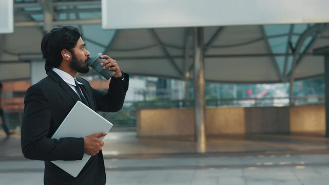 Side view of classy man carrying digital gadget and reusable cup for drink while listening radio through wireless earphones. Motivated company manager pacing on way to office building by city.