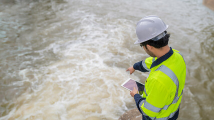 A engineering doing his checking routine. He is wearing hard hat and engineer uniform.Standing by the rail by the dam.Monitor water levels from the heavy rain that has been falling for several days.