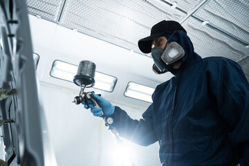 Mechanic from vehicle maintenance service is spreading paint on car using dark spray and paint gun. Professional worker wearing protective gloves is painting automobile.