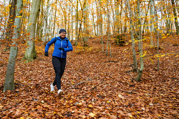 Fototapeta premium Middle-aged woman dressed sporty in blue jacket and black pants, cap and glasses running downhill in forest full of fallen leaves in autumn weather. Front view