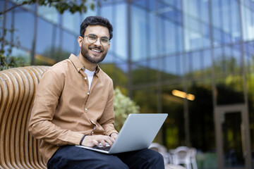 Young man with beard and glasses sits on bench outside modern building, using laptop. He is...