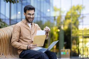 Smiling man sitting on bench reading letter from envelope with laptop on lap. Outdoor scene conveys...