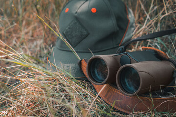 Outdoor gear featuring binoculars and a green cap lying in dry grass, symbolizing a nature hike, camping, or hunting scene, perfect for themes of exploration, adventure, or wildlife observation © solidmaks