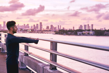 Man, arm stretch and city skyline on pier with sunrise, athlete and fitness break for training in the morning. Exercise, urban and confidence by the sea ready for run and marathon at dawn in New York