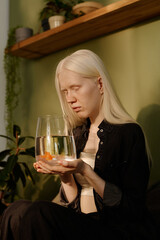Vertical shot of gen Z girl with albinism holding small aquarium in hands and looking at her pet goldfish