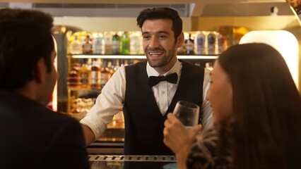Focusing smiling smart bartender serving gin fizz or tonic soda at luxurious bar, engaging lively conversation with VIP customer couple at back view in atmosphere at lighting warm nightclub. Vinosity.